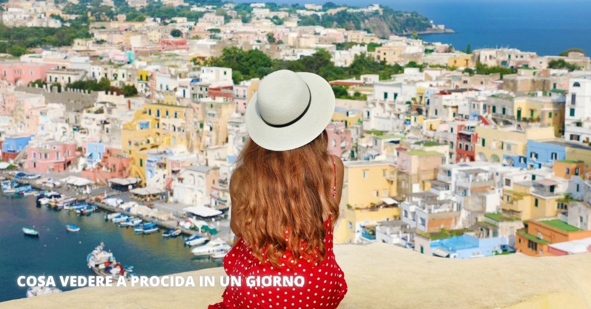 Travels in italy. back view of beautiful girl with hat sitting on wall looking at stunning panoramic view of procida island, naples, italy. procida is italian capital of culture 2022